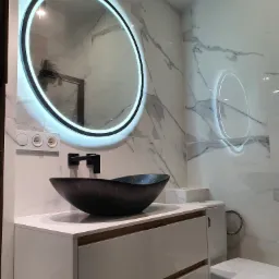 Nowoczesna łazienka z umywalką w kształcie misy, podświetlanym okrągłym lustrem i marmurowymi płytkami na ścianach.