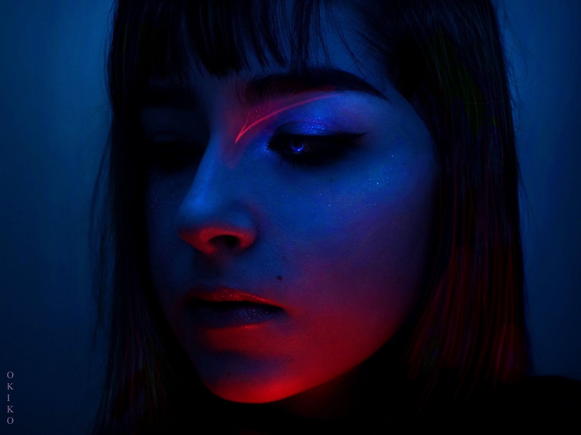 Portret kobiety w chłodnym, niebieskim oświetleniu z geometrycznym, czerwonym akcentem makijażu nad brwią i błyszczącym makijażem oczu.
