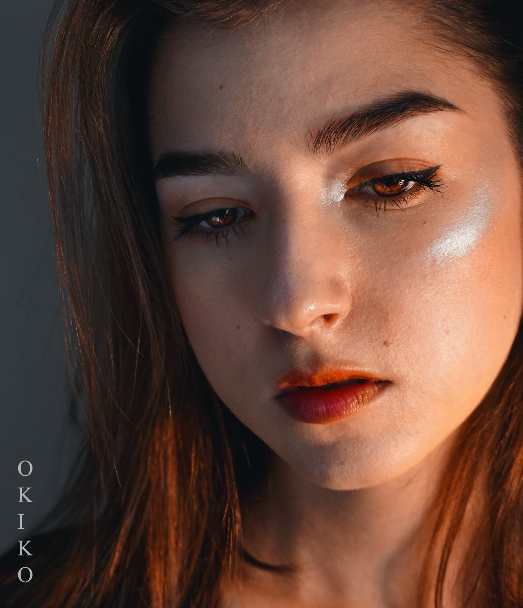 Portret kobiety z makijażem, podkreślone oczy eyelinerem, pomarańczowe usta, oświetlenie boczne tworzące cienie na twarzy, widoczne logo OKIKO po lewej stronie.