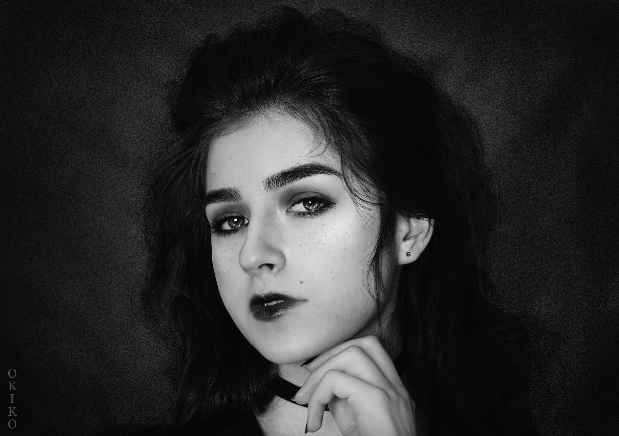 Portret młodej kobiety z ciemnymi włosami i makijażem w stylu vintage, czarno-białe zdjęcie z delikatnym ziarnem, choker na szyi i dłoń pod brodą.