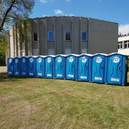 Rząd niebieskich, przenośnych toalet ustawionych na trawie przed budynkiem z szarymi kolumnami i oknami, pod błękitnym niebem.