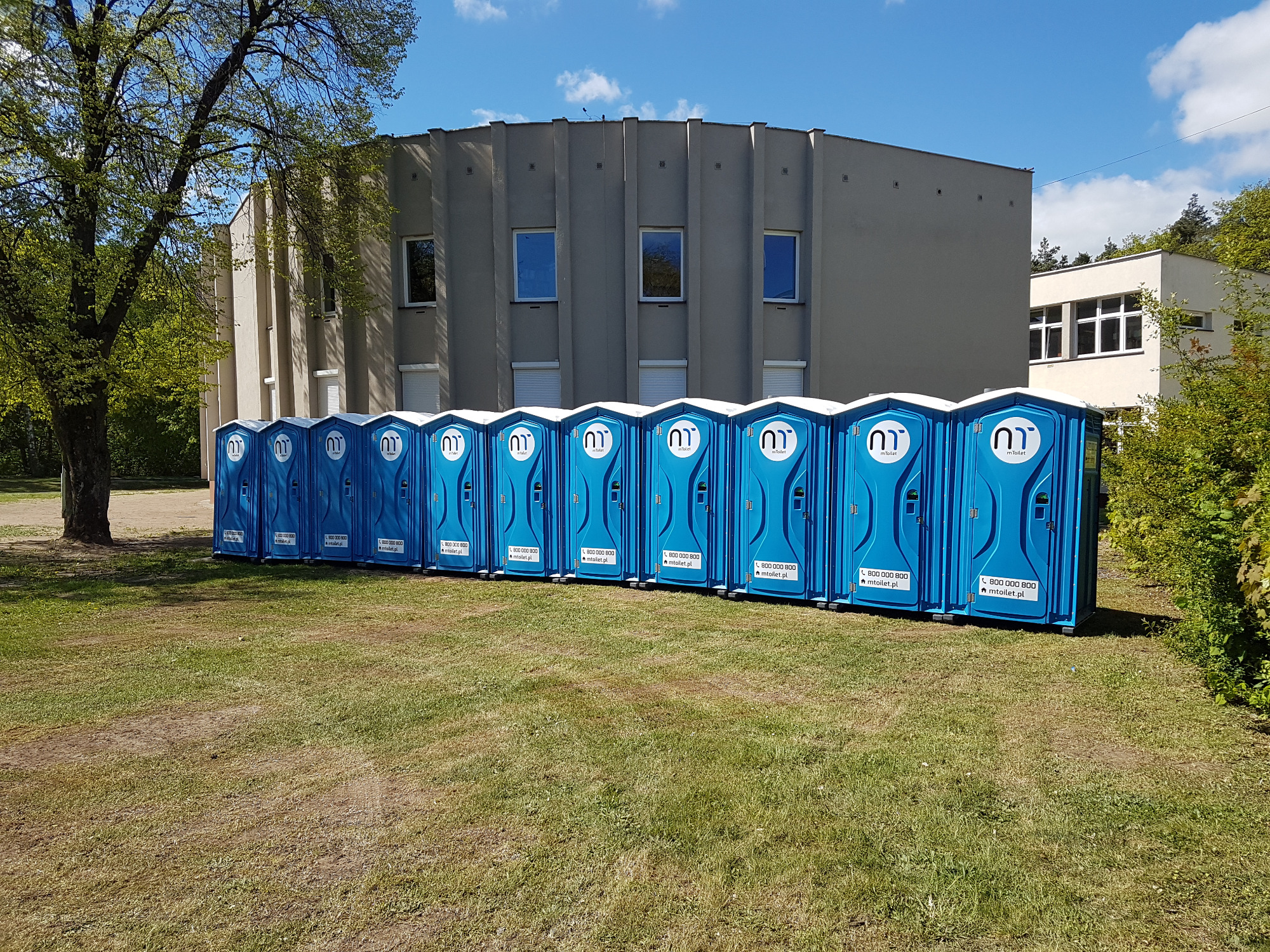 Rząd niebieskich, przenośnych toalet ustawionych na trawie przed budynkiem z szarymi kolumnami i oknami, pod błękitnym niebem.