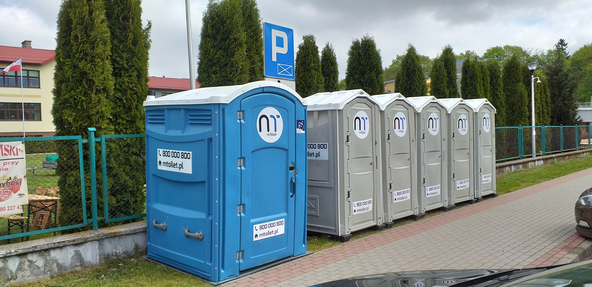 Rząd przenośnych toalet w kolorach niebieskim i szarym, ustawionych na chodniku, z logo i numerem telefonu firmy.
