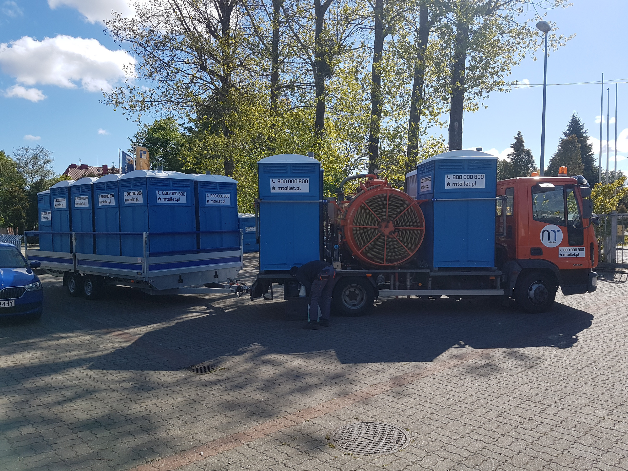 Ciężarówka z przyczepą przewożąca przenośne toalety, na boku widoczny pomarańczowy, spiralny element czyszczący kanalizację.