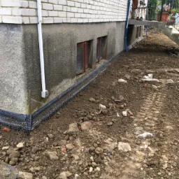 Instalacja drenażu opaskowego przy fundamentach budynku z cegły i betonu, widoczne rury odprowadzające wodę i mata drenażowa, ziemia po wykopie.