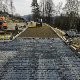 Częściowo ułożona, szara kostka brukowa na podjeździe w Lesku, z widocznym równaniem podłoża i ciężkim sprzętem w tle, w pochmurny dzień.