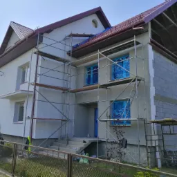 Dwupiętrowy dom w trakcie prac elewacyjnych, z oknami zabezpieczonymi folią, otoczony rusztowaniami i częściowo otynkowany, z widoczną ścianą z bloczków betonowych.