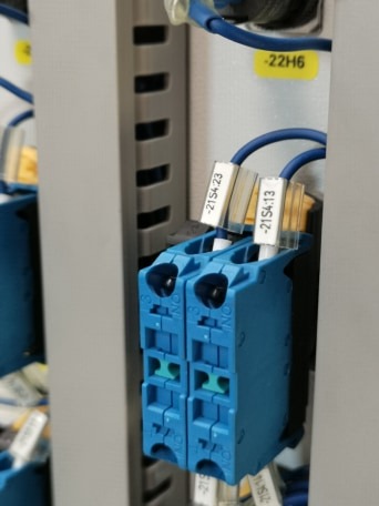 Detale podłączenia elektrycznego: dwa niebieskie bloki zaciskowe z przewodami oznaczonymi etykietami -21S4.23 i -21S4.13, zamontowane w szarej szafie sterowniczej.