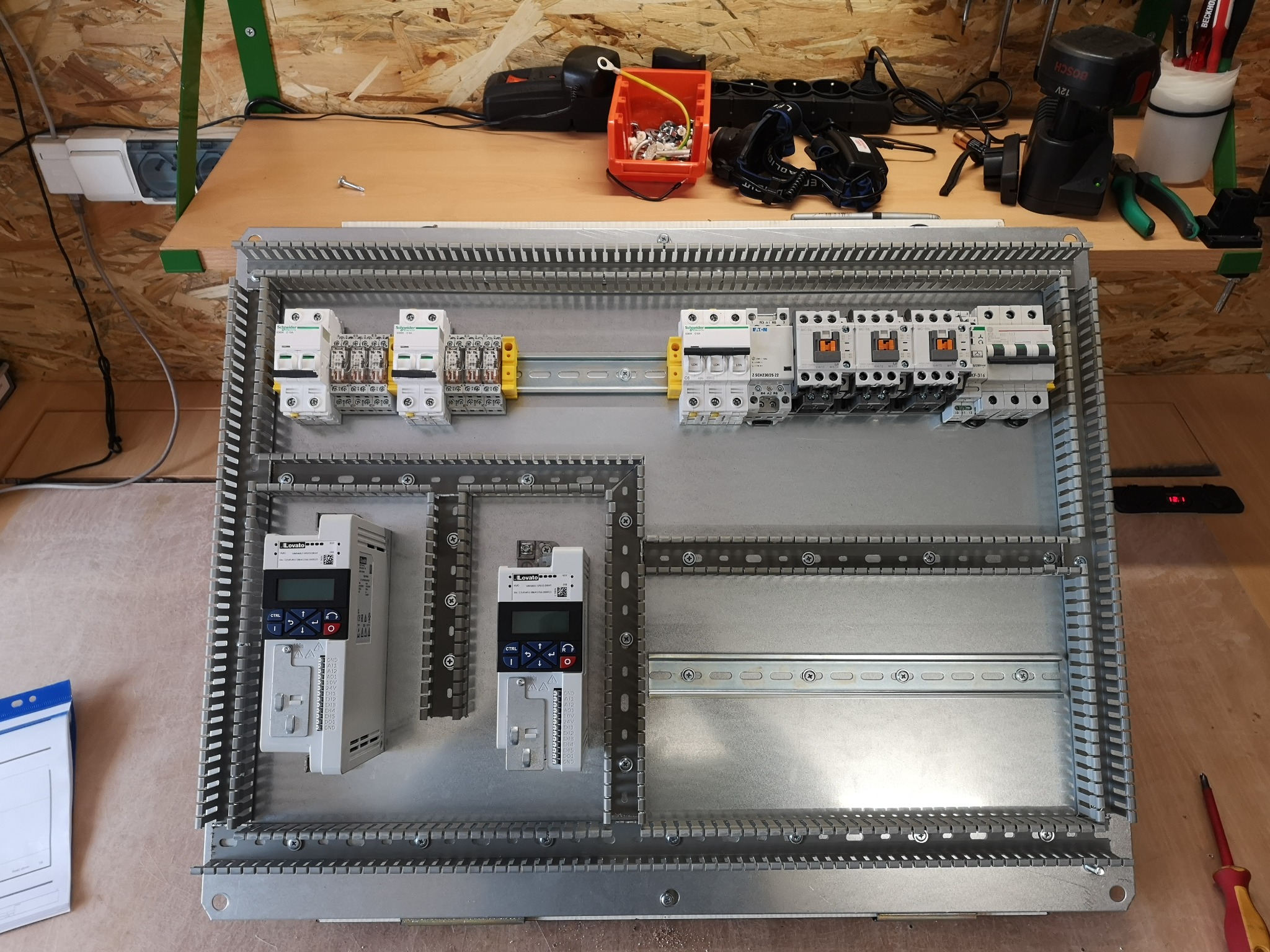 Rozdzielnica elektryczna z zamontowanymi zabezpieczeniami, stycznikami i falownikami, widok z góry na stole warsztatowym z narzędziami.