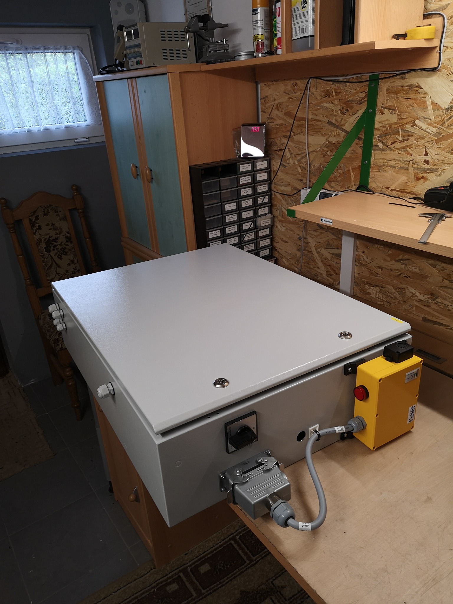 Szara skrzynka sterownicza z żółtym przyciskiem awaryjnym i podłączonymi przewodami, umieszczona na stole warsztatowym w pomieszczeniu z widocznym wyposażeniem warsztatowym.