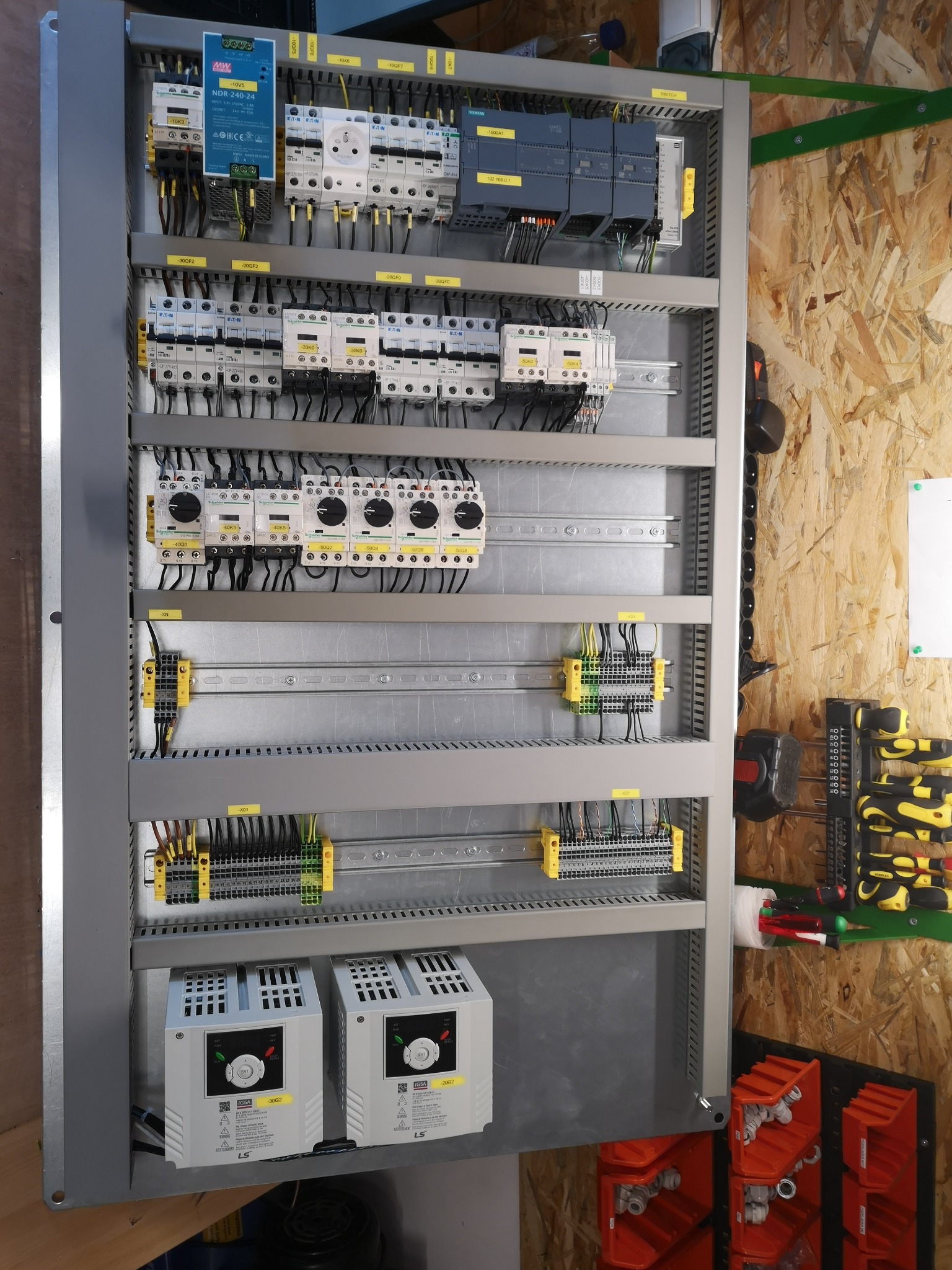 Szara szafa sterownicza z zamontowanymi elementami elektrycznymi, w tym stycznikami, zasilaczami i falownikami LS, widoczne okablowanie i oznaczenia, obok ściana z płyty OSB z narzędziami.