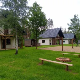 Kompleks domków letniskowych z drewnianymi balkonami, placem zabaw i miejscem na ognisko z drewnianymi ławkami, otoczony zielenią i drzewami.