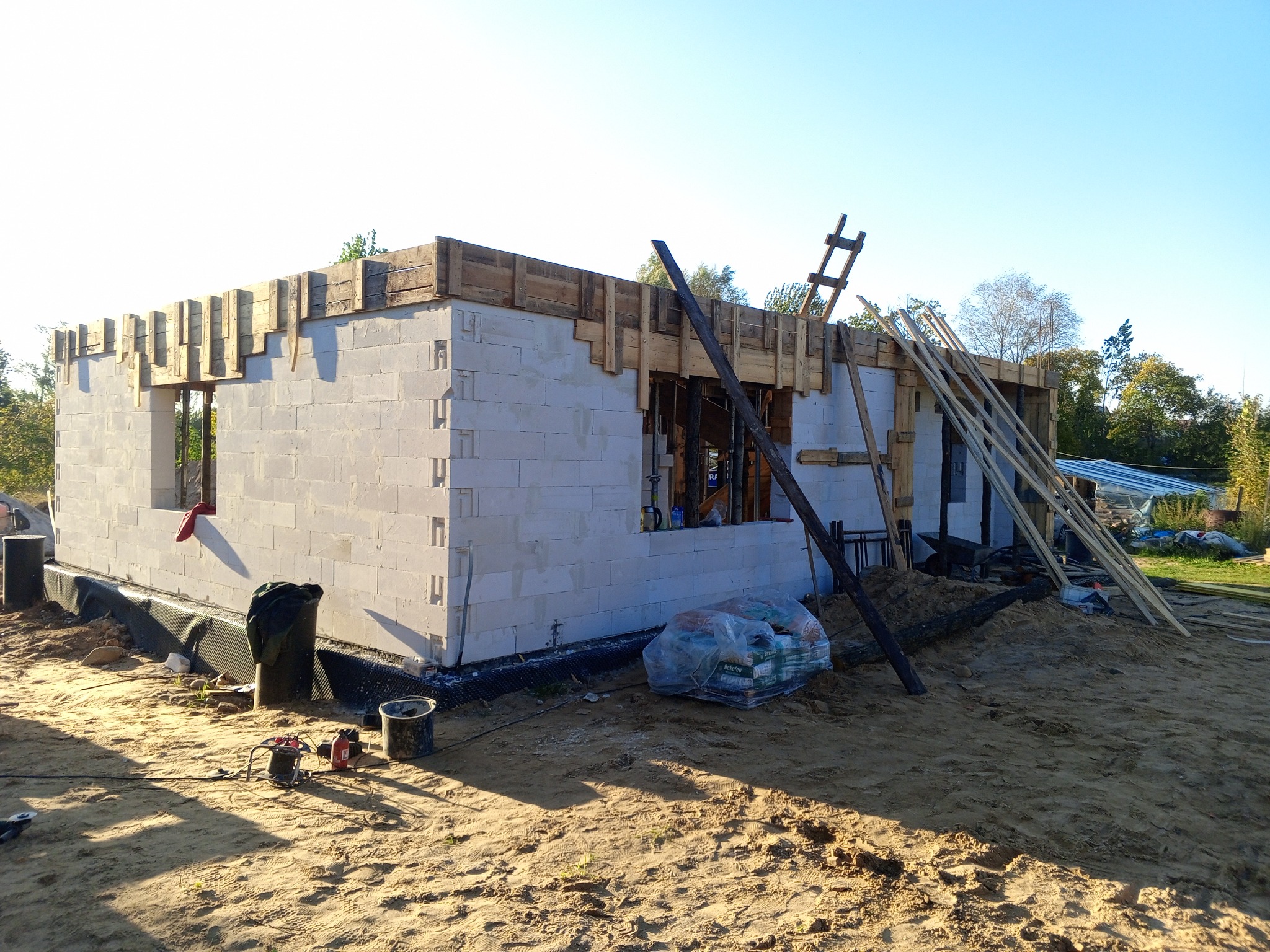 Budowa domu jednorodzinnego: ściany z betonu komórkowego w trakcie wznoszenia, drewniane szalunki nad otworami okiennymi, czarna izolacja fundamentów, narzędzia budowlane na piaszczystym podłożu.