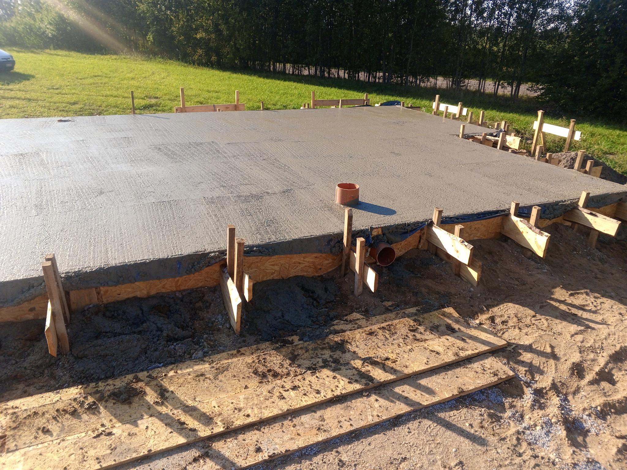 Wylany betonowy fundament z drewnianym szalunkiem i rurą kanalizacyjną, widok z góry na tle trawnika.