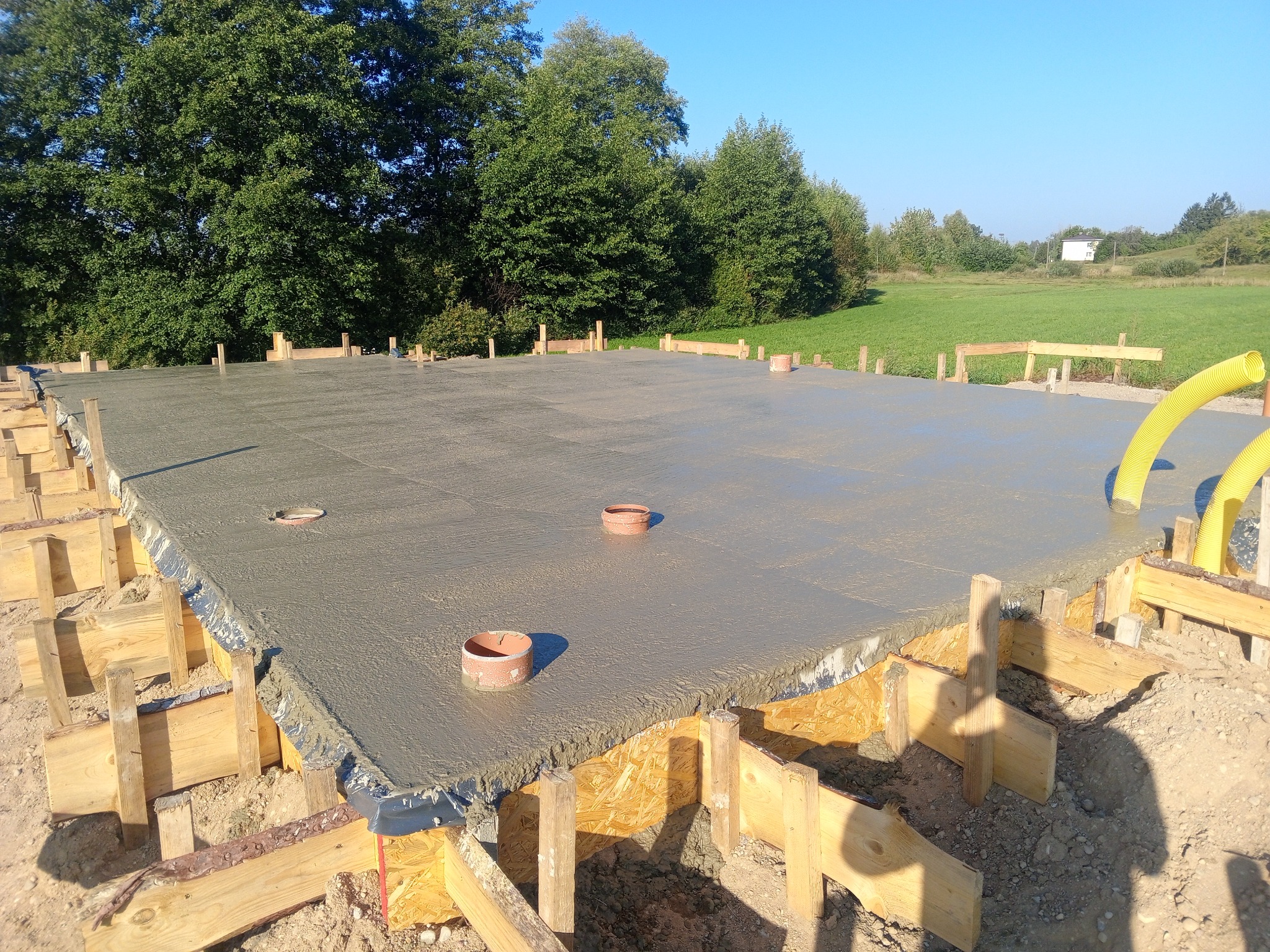 Wylany fundament płyty betonowej z widocznymi rurami kanalizacyjnymi, szalunkiem z desek i folią izolacyjną w słoneczny dzień.