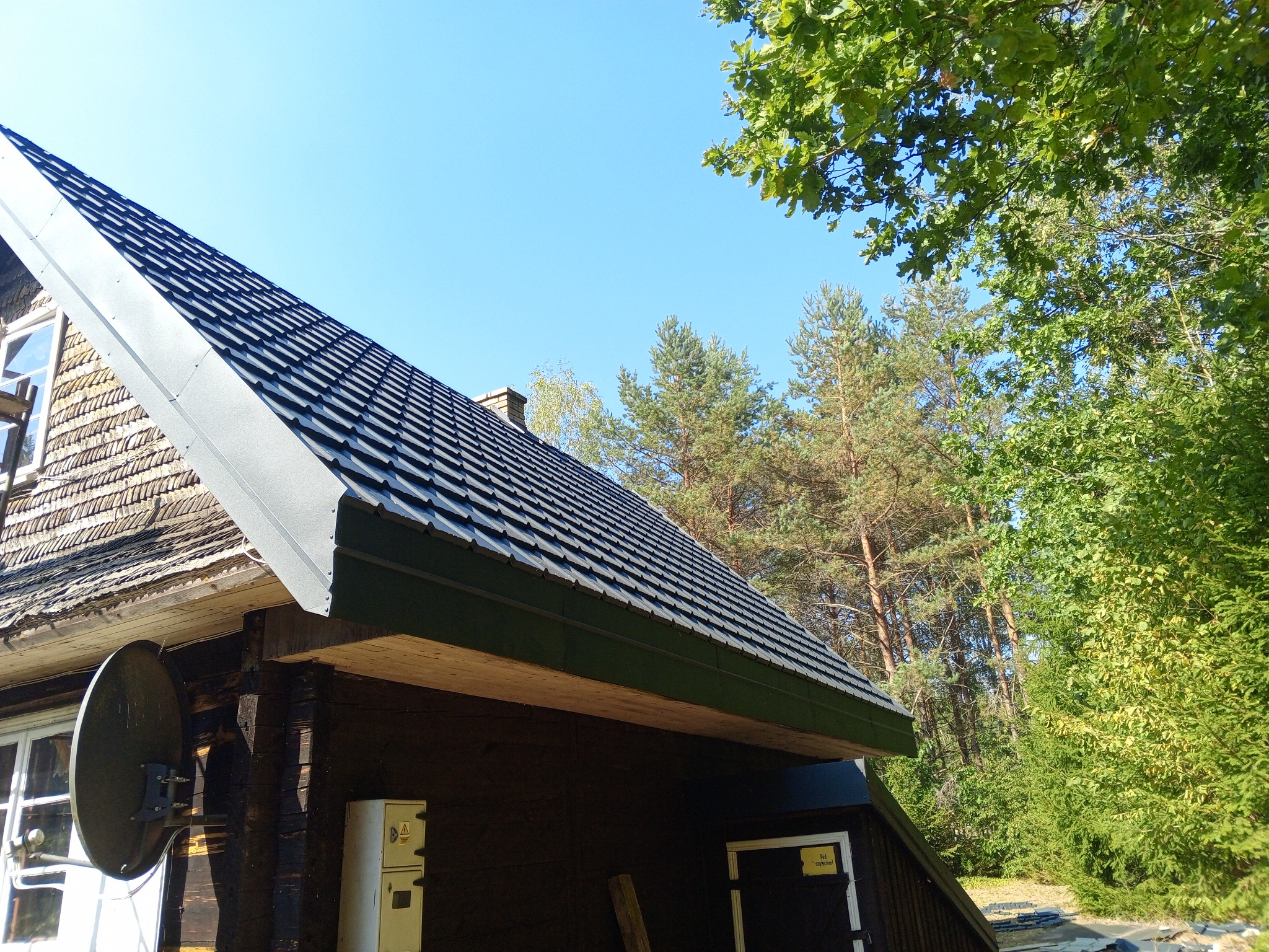 Wykończony dach z czarnej dachówki na drewnianym domu z widoczną anteną satelitarną i fragmentem lasu w tle.