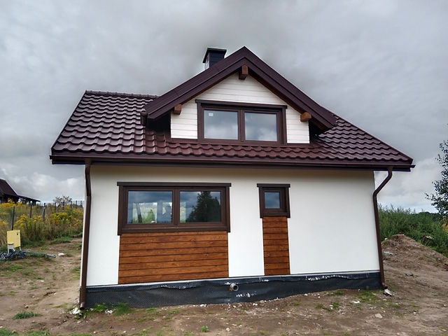 Ocieplony dom jednorodzinny z brązową dachówką i drewnianą boazerią na dolnej części elewacji, widoczne okna z brązowymi ramami i rynny w tym samym kolorze, całość na tle pochmurnego nieba.