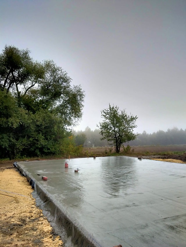 Wylana, świeża płyta fundamentowa na gruncie w pochmurny dzień, zabezpieczona folią, z widocznymi poziomnicami i otaczającą zielenią.