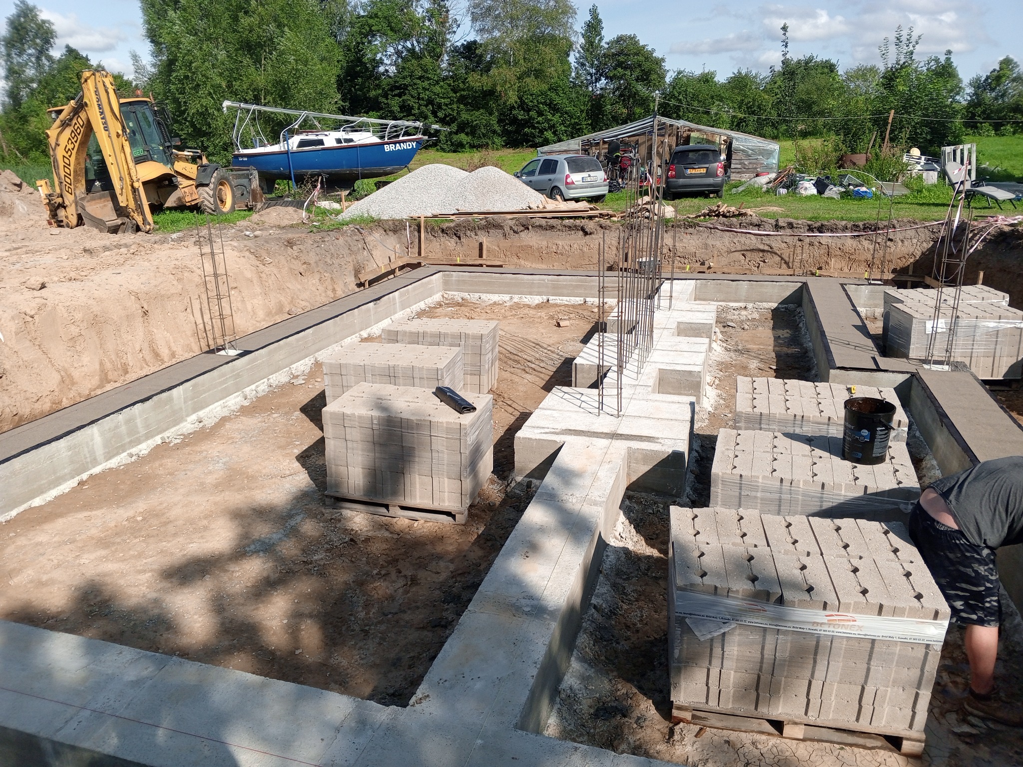 Wykop pod fundamenty domu jednorodzinnego z widoczną zbrojoną ławą fundamentową, ułożonymi bloczkami betonowymi, koparką i jachtem w tle.