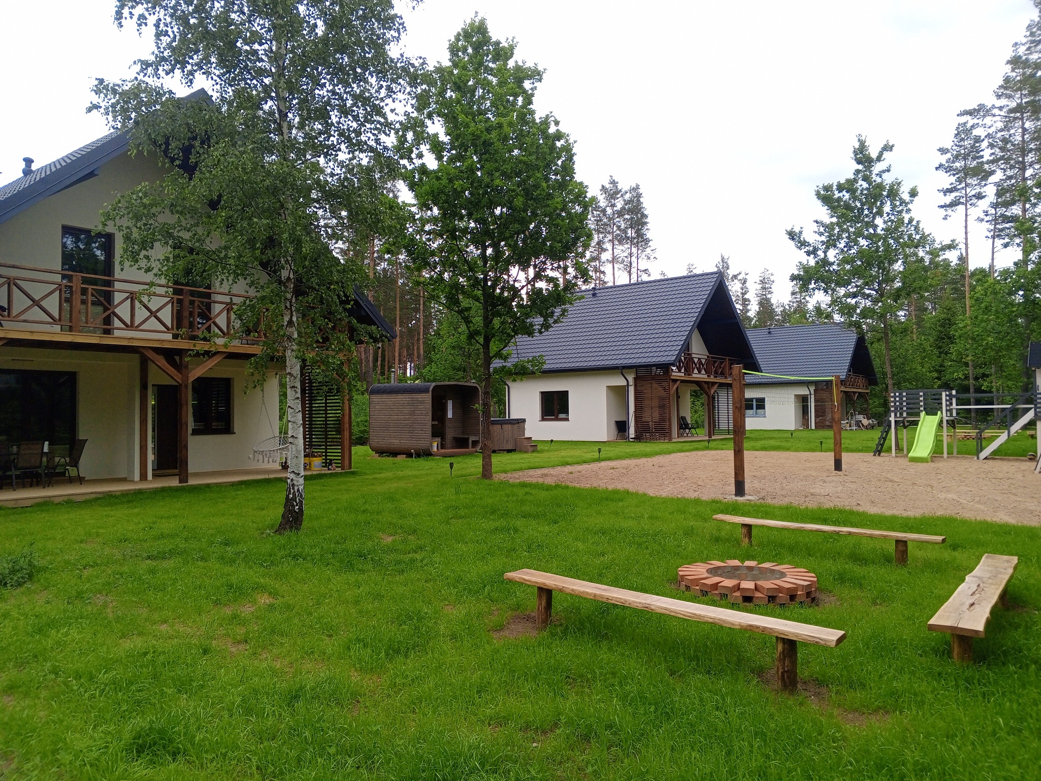 Kompleks domków letniskowych z drewnianymi balkonami, placem zabaw i miejscem na ognisko z drewnianymi ławkami, otoczony zielenią i drzewami.