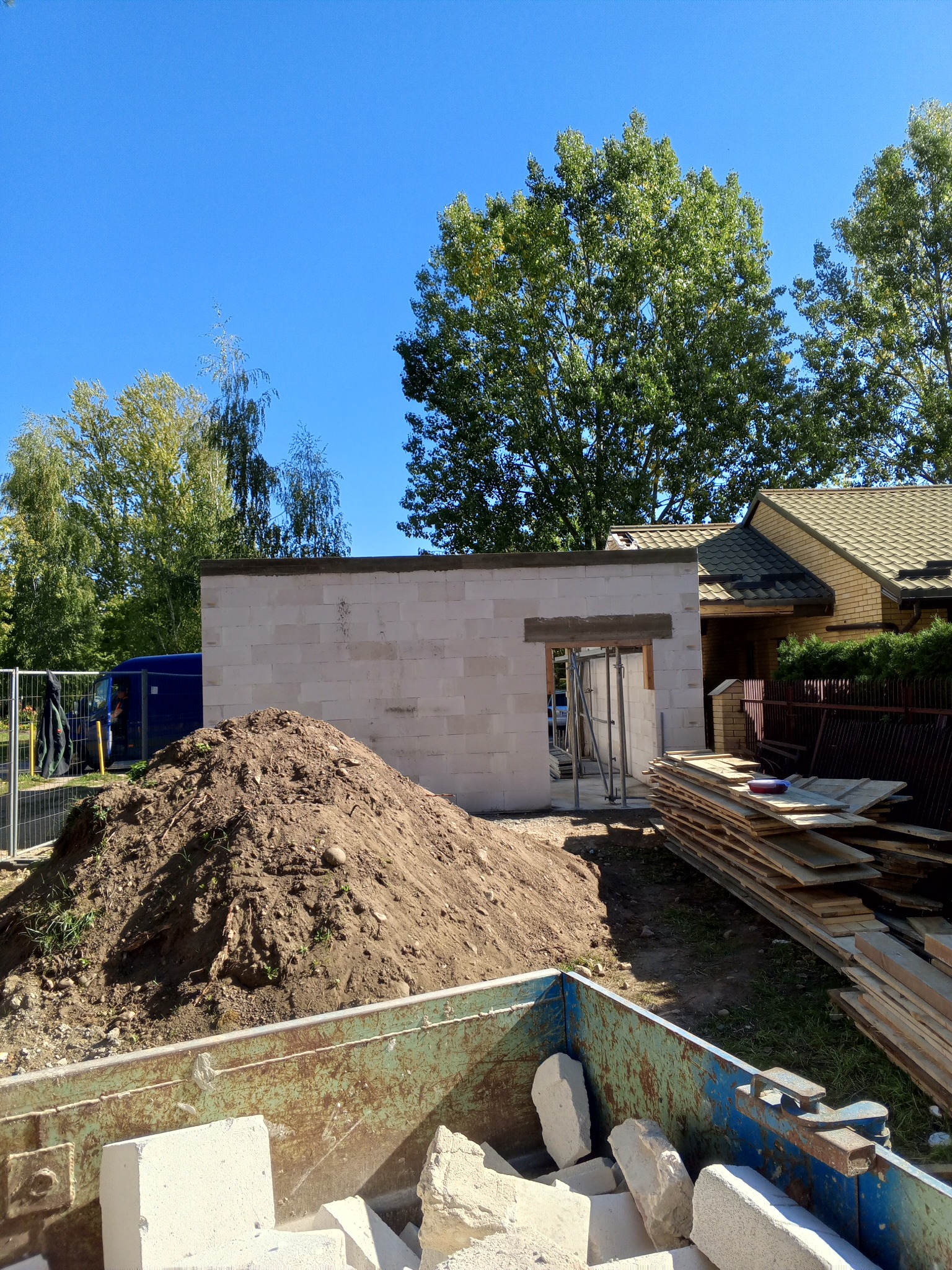 Wznoszenie ściany z bloczków betonowych, widoczny otwór okienny, sterta ziemi, kontener z gruzem i stos desek na placu budowy w słoneczny dzień.