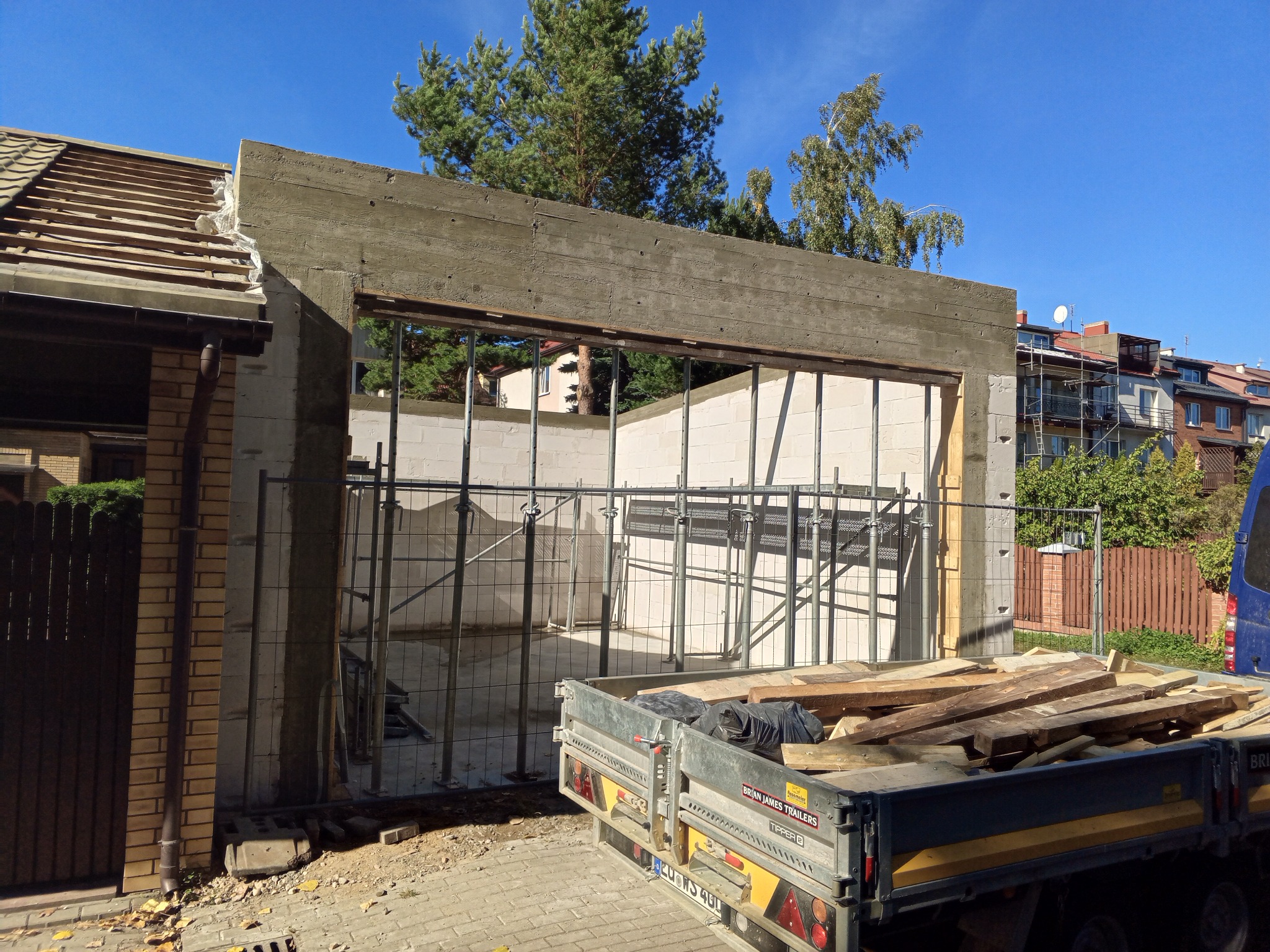 Wznoszenie betonowego fundamentu z widocznym zbrojeniem i deskowaniem, na tle częściowo zdjętego dachu oraz przyczepy z materiałami budowlanymi.