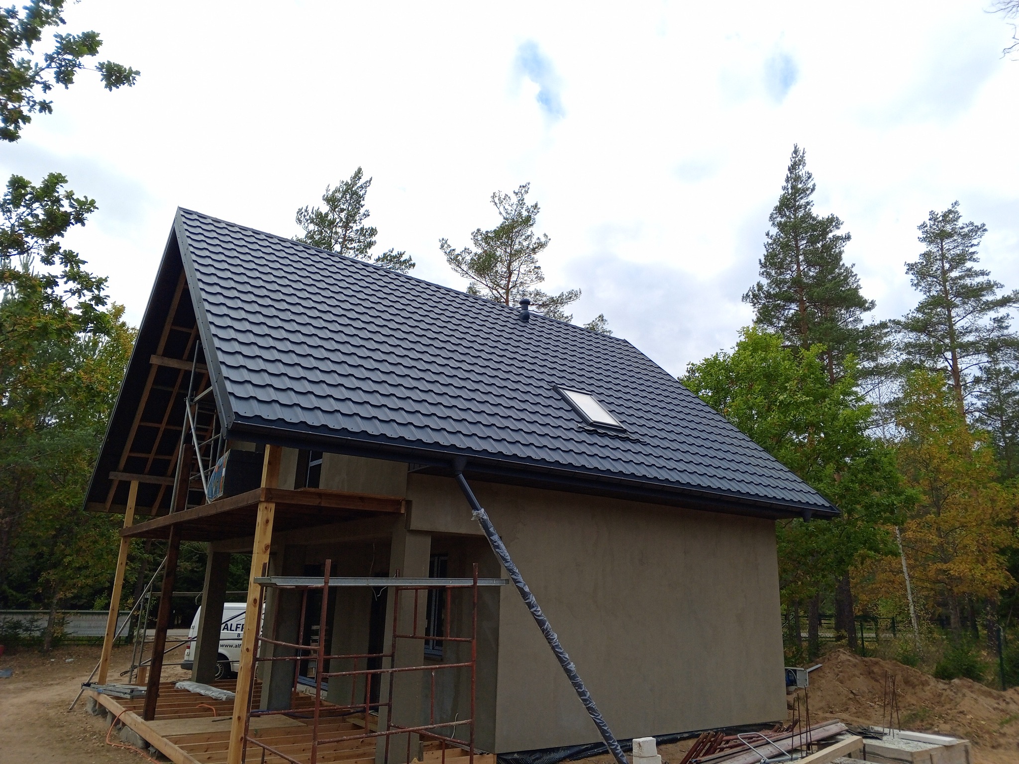 Dom w trakcie budowy z nowym, grafitowym dachem z blachodachówki i widoczną więźbą dachową, częściowo otynkowany, z rusztowaniem i materiałami budowlanymi na posesji.