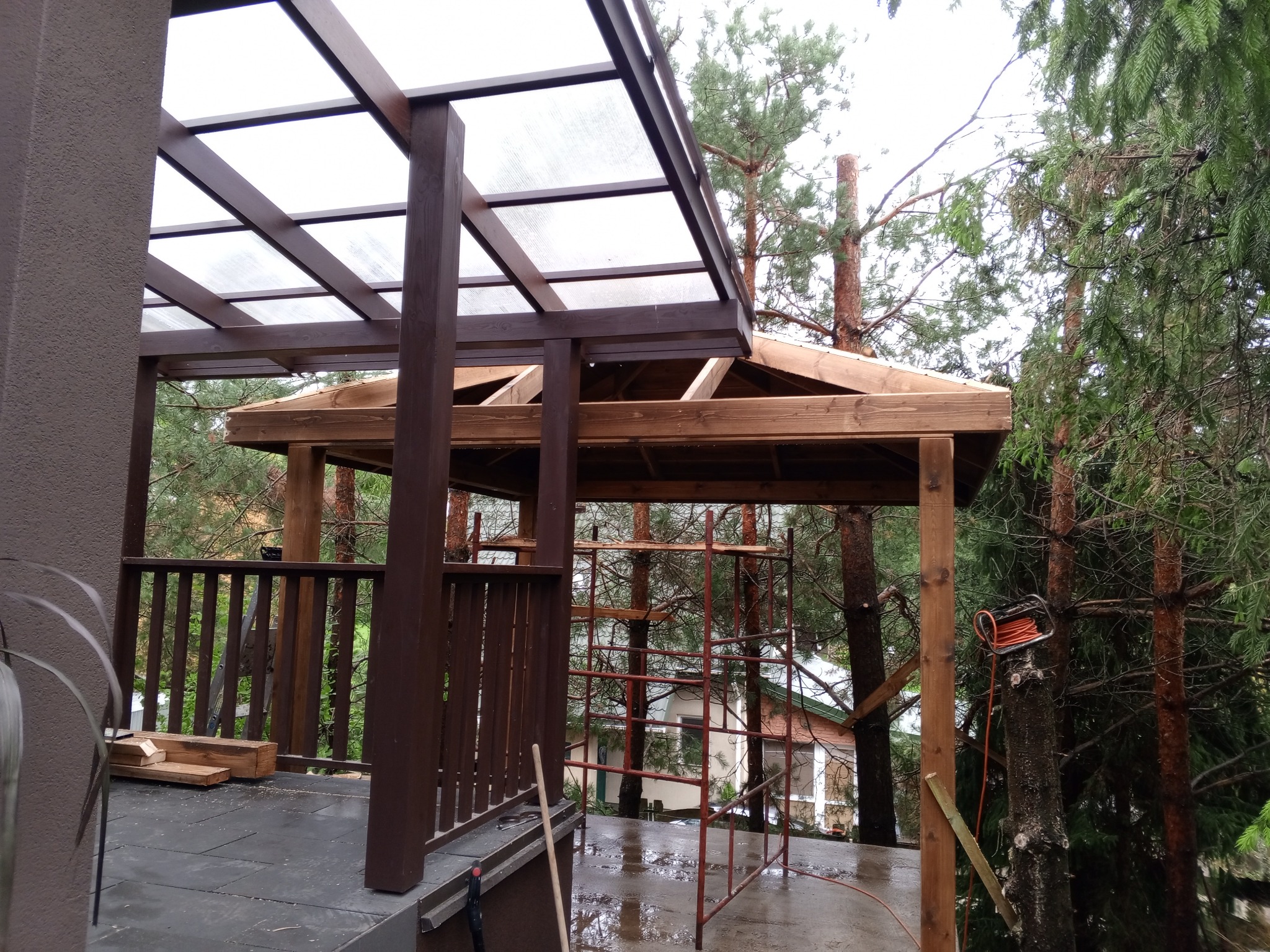 Drewniana konstrukcja z przezroczystym zadaszeniem w trakcie budowy, widoczne rusztowanie, kabel elektryczny zwisający z drzewa, otoczenie leśne i dom w tle.