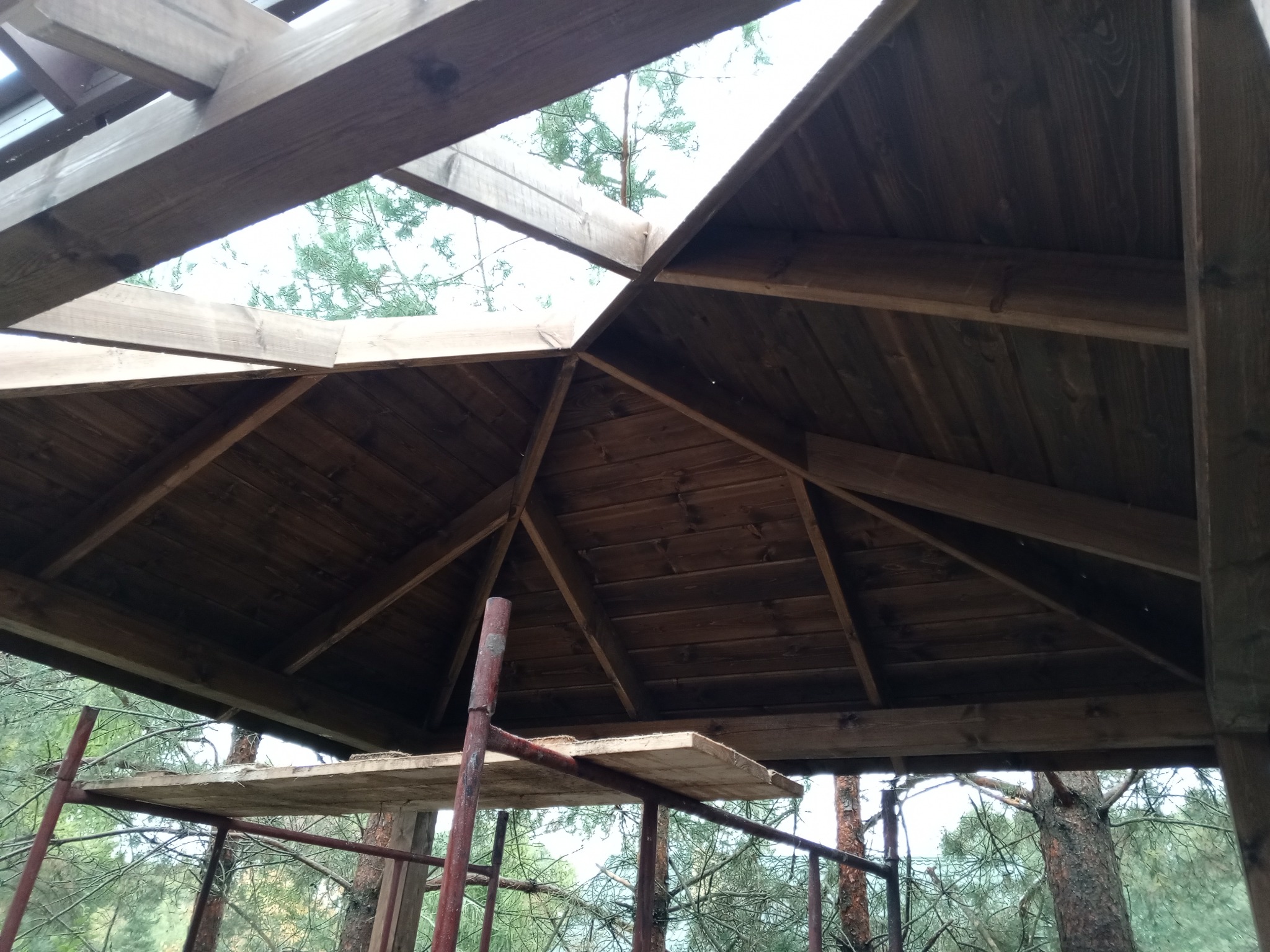 Wnętrze drewnianej konstrukcji dachu altany w trakcie budowy, widoczne rusztowanie i drzewa w tle.