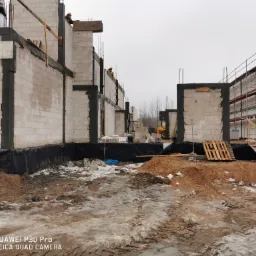 Wznoszenie ścian z bloczków na budowie domu, widoczni pracownicy w kaskach, teren budowy z błotem i resztkami śniegu, w tle żółta koparka.