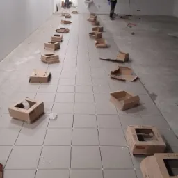 Układanie szarych płytek podłogowych w długim pomieszczeniu, z kartonowymi pudełkami po płytkach rozstawionymi wzdłuż ułożonej części, widoczni pracownicy w odblaskowych kamizelkach w tle.