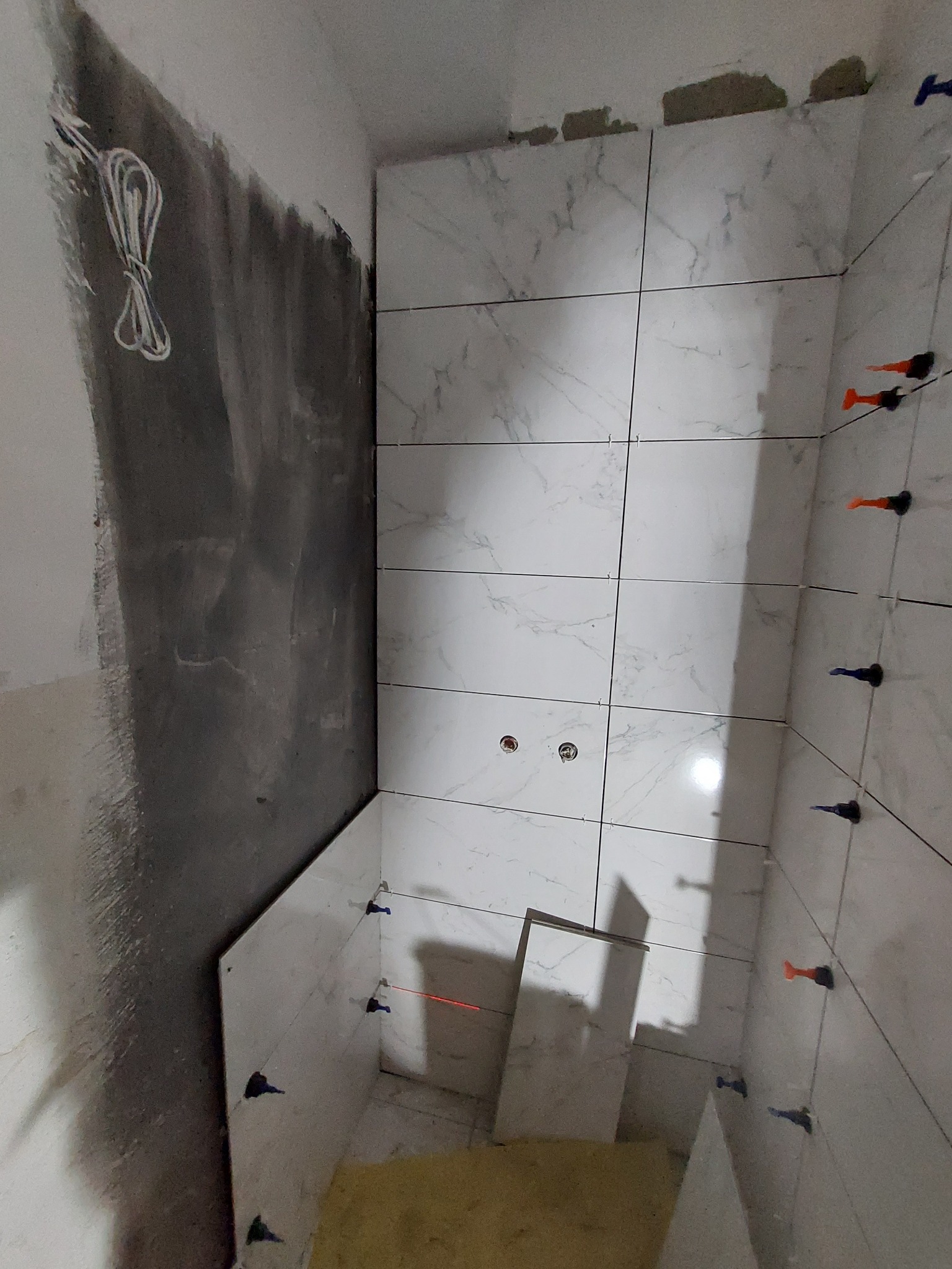 Wnętrze łazienki w trakcie układania białych płytek imitujących marmur, widoczne krzyżyki dystansowe, laserowy poziom, ściana surowa przygotowana pod płytki, wystające przewody.