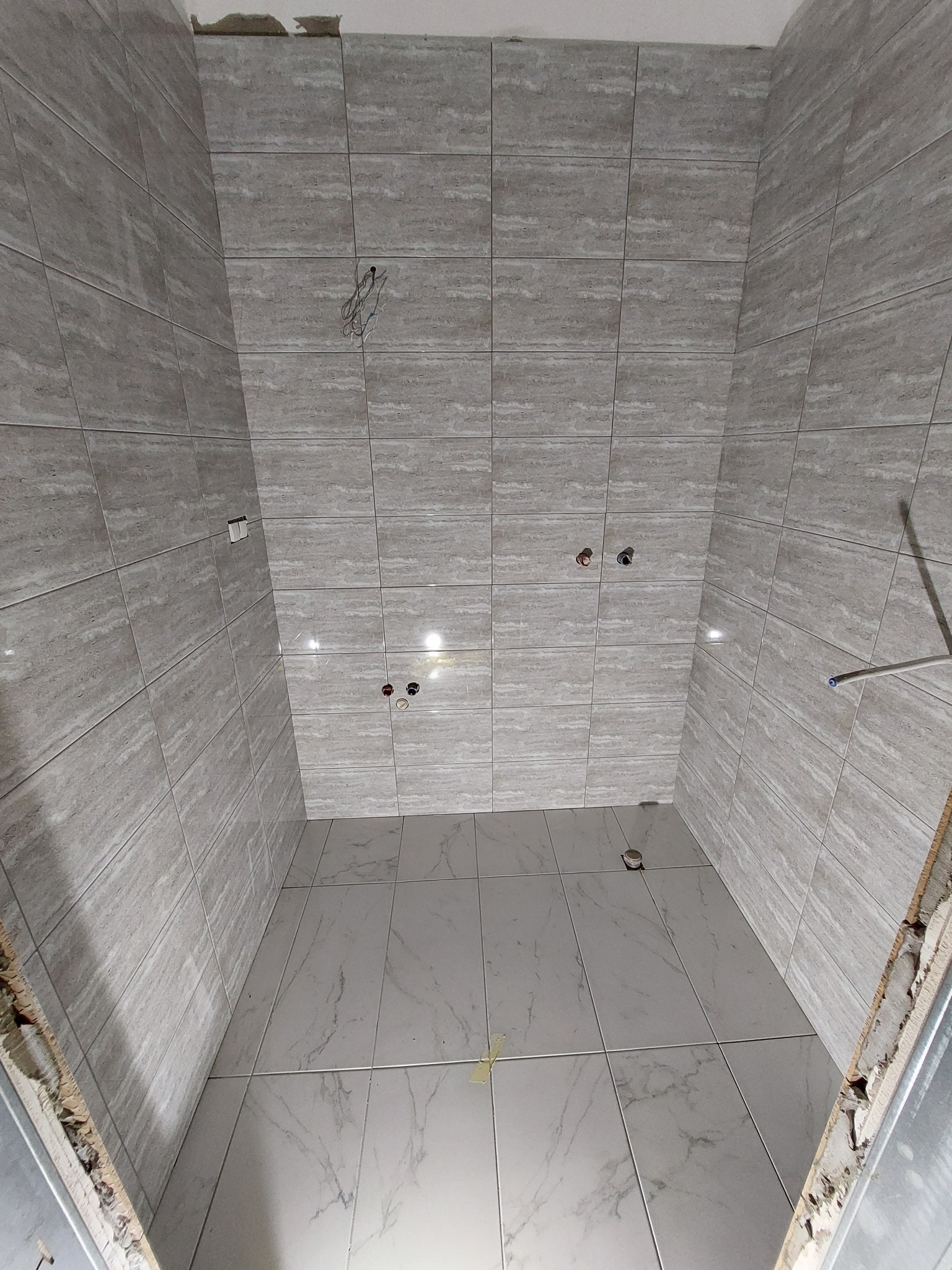 Płytki imitujące kamień na ścianach i marmur na podłodze w trakcie montażu w łazience, widoczne wystające przewody i rury instalacji wodnej.