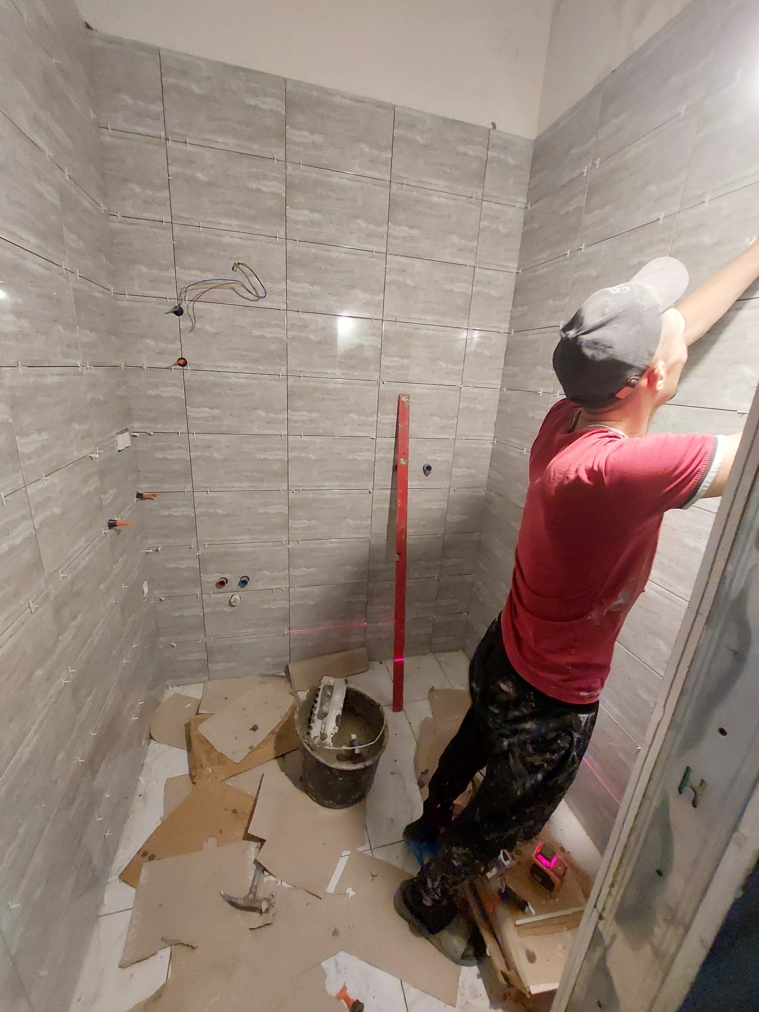 Układanie szarych płytek w łazience z użyciem poziomicy laserowej, widoczne krzyżyki dystansowe, wiadro z zaprawą i narzędzia na podłodze.