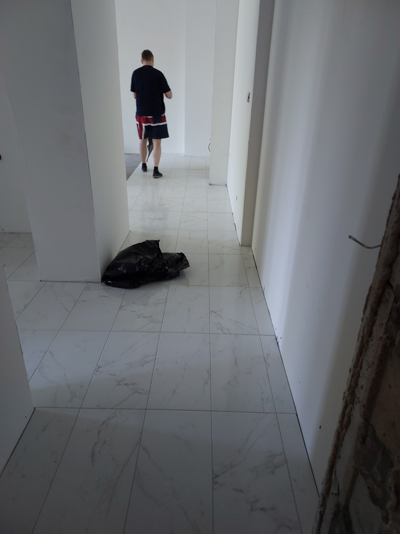 Korytarz po ułożeniu białych płytek podłogowych imitujących marmur, widoczny czarny worek na śmieci i osoba oddalająca się w głąb pomieszczenia.