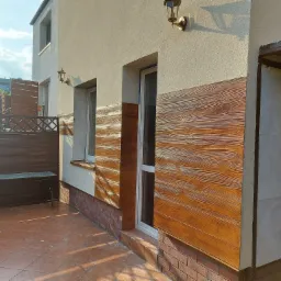 Wykończona elewacja domu z panelami imitującymi drewno, widoczne okna, drzwi i fragment tarasu z kostki brukowej.
