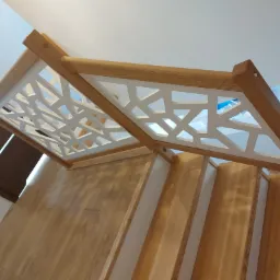 Drewniane schody z nowoczesną, ażurową balustradą w geometryczne wzory, wykończone w jasnym drewnie, widok z dołu.