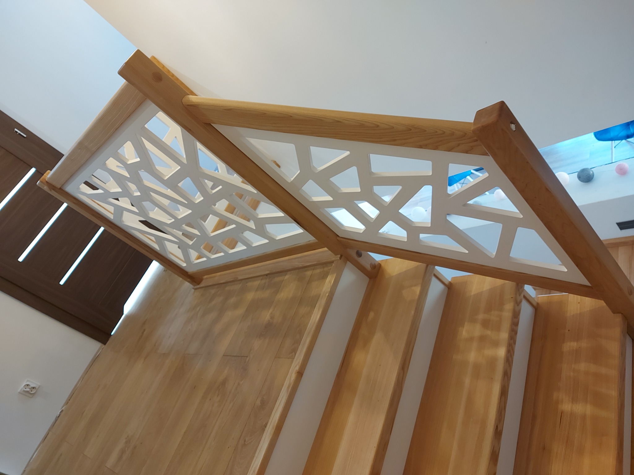 Drewniane schody z nowoczesną, ażurową balustradą w geometryczne wzory, wykończone w jasnym drewnie, widok z dołu.