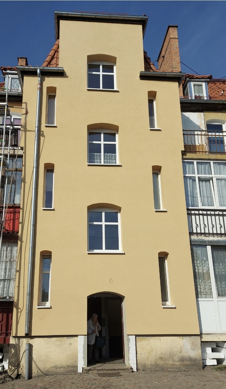 Żółta elewacja kamienicy z białymi oknami, widoczna osoba w wejściu, fragment rusztowania po lewej stronie.