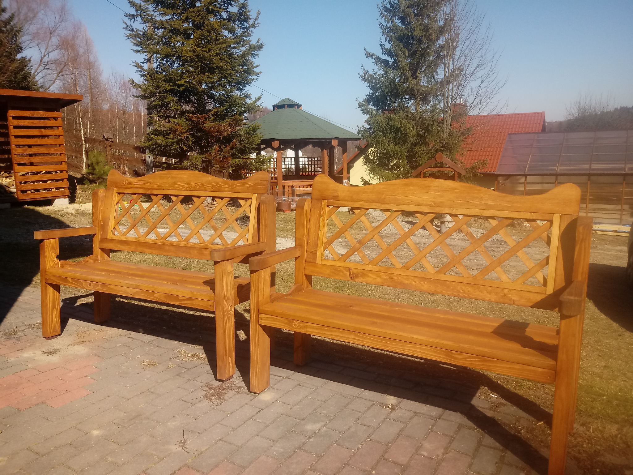 Dwie identyczne, świeżo polakierowane drewniane ławki ogrodowe z kratownicą na oparciu, ustawione na kostce brukowej przed altaną ogrodową z zielonym dachem.