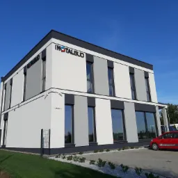 Nowoczesny budynek biurowy z logo 'INSTALBUD' na fasadzie, biało-szara kolorystyka, monitoring, czerwony samochód firmowy zaparkowany przed budynkiem, adres 'Lakowa 18' widoczny na rogu budynku.