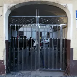 Metalowa brama wjazdowa z ozdobnymi elementami i kulami, widoczna z zewnątrz przez kamienną arkadę, w tle wnętrze warsztatu z samochodami.