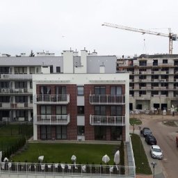 OKSANBUD Sp. z o.o. - Nowoczesne budynki mieszkalne z balkonami, w tle budowa z dźwigiem. Widok z góry na osiedle z zielenią i zaparkowanymi autami. Szara, pochmurna pogoda.
