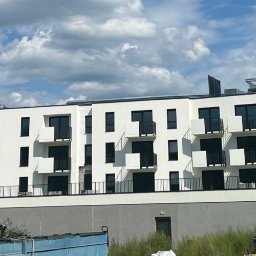 OKSANBUD Sp. z o.o. - Nowoczesny, trzypiętrowy budynek apartamentowy z białymi ścianami i czarnymi balkonami na tle błękitnego nieba z obłokami. Widoczne elementy placu budowy.