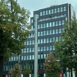 OKSANBUD Sp. z o.o. - Nowoczesny, ciemny budynek biurowy z licznymi oknami, widziany z poziomu ulicy, z zielenią drzew po bokach. Widoczny szyld Santander na parterze.