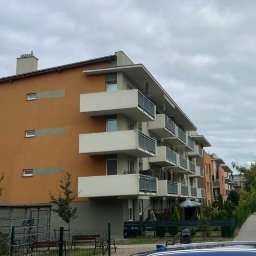 OKSANBUD Sp. z o.o. - Nowoczesny, pomarańczowy budynek wielorodzinny z balkonami, widziany z perspektywy ulicy. Widoczne drzewa i chodnik. Pochmurne niebo w tle.