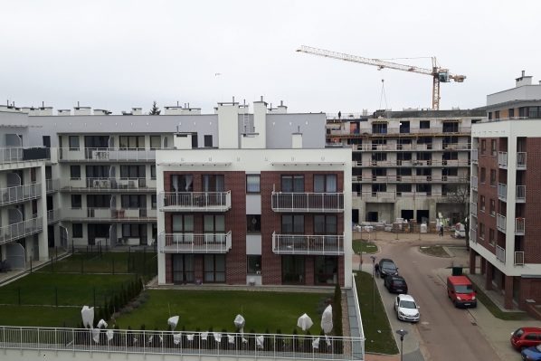 Nowoczesne budynki mieszkalne z balkonami, w tle budowa z dźwigiem. Widok z góry na osiedle z zielenią i zaparkowanymi autami. Szara, pochmurna pogoda.