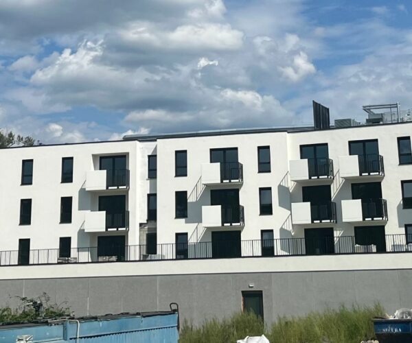 Nowoczesny, trzypiętrowy budynek apartamentowy z białymi ścianami i czarnymi balkonami na tle błękitnego nieba z obłokami. Widoczne elementy placu budowy.