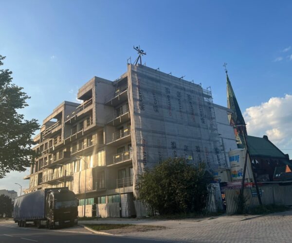 Wznoszony budynek żelbetowy z widoczną konstrukcją, zabezpieczony siatką, na tle kościoła i błękitnego nieba. Na pierwszym planie zaparkowana ciężarówka.