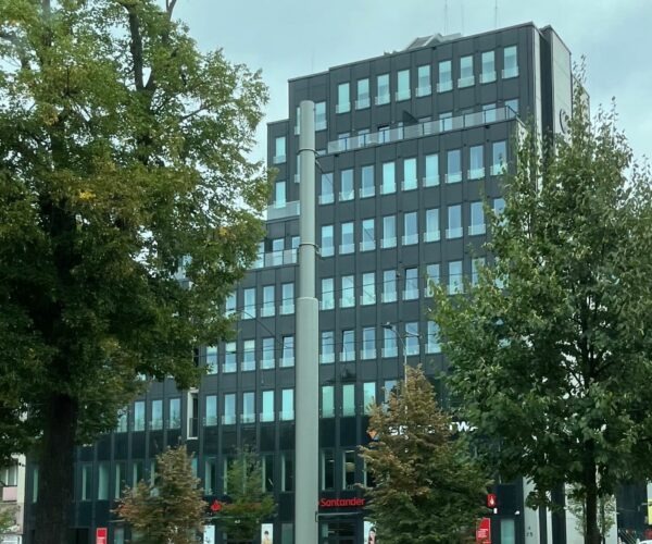 Nowoczesny, ciemny budynek biurowy z licznymi oknami, widziany z poziomu ulicy, z zielenią drzew po bokach. Widoczny szyld Santander na parterze.